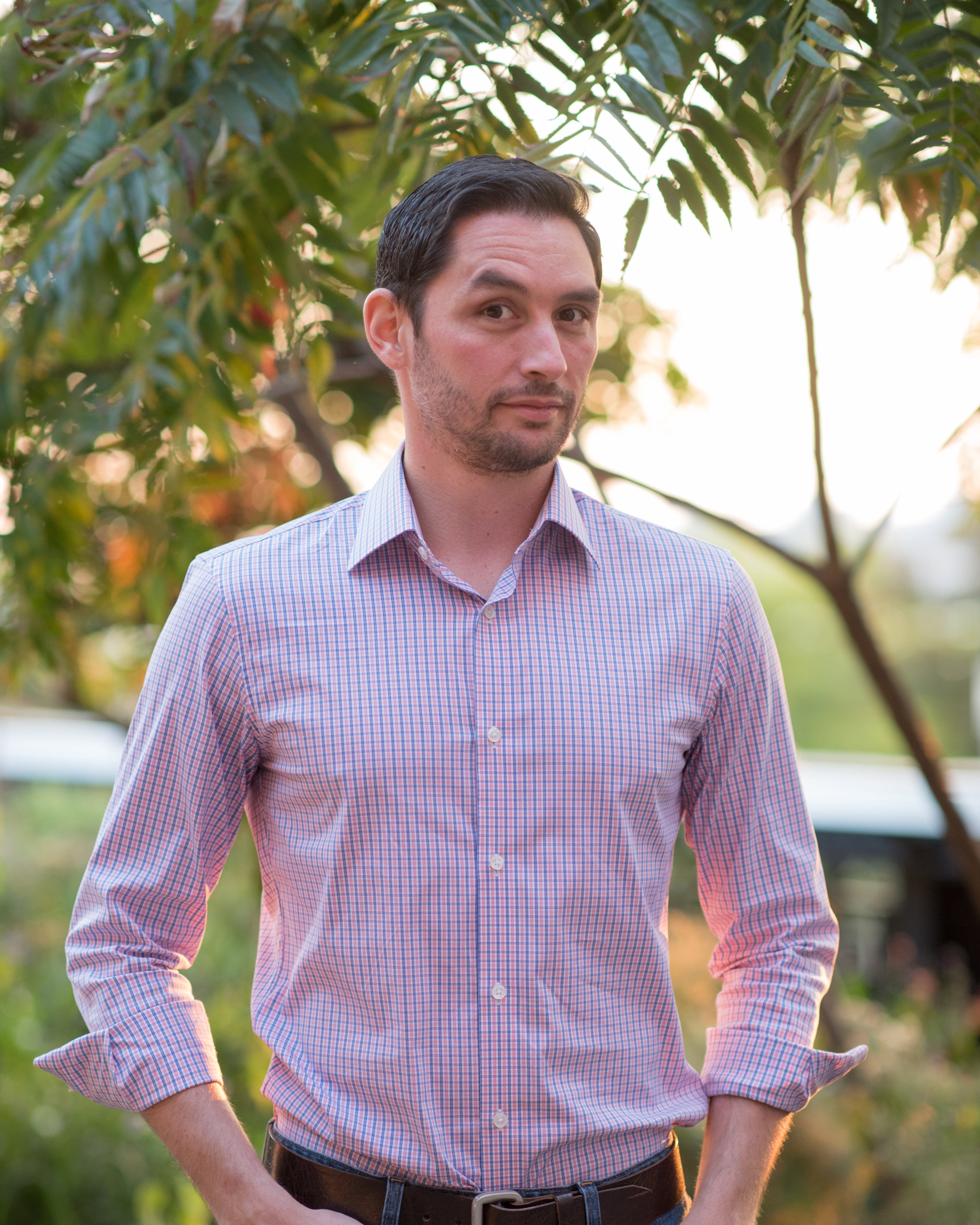 Man wearing a pink checkered shirt and blue jeans standing outdoors with greenery in the background